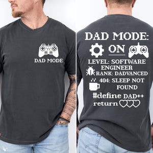May include: Dark gray t-shirt with white text and graphics. The front features a game controller and the words "DAD MODE." The back displays "DAD MODE: ON" with a gear, controller, and other text, including "SOFTWARE ENGINEER" and "#define DAD++."