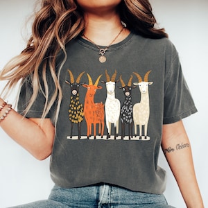 May include: Dark gray t-shirt featuring a graphic of five stylized goats in various colors: black, orange, white, and cream. The goats stand side-by-side on a white line. The shirt is a comfortable, casual style.