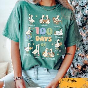 May include: Light green t-shirt with a graphic featuring cartoon geese engaged in various activities, including reading and creating. The text "100 Days of" is prominently displayed. The shirt is a relaxed fit.
