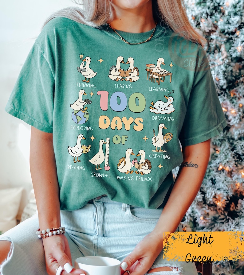 May include: Light green t-shirt with a graphic featuring cartoon geese engaged in various activities, including reading and creating. The text "100 Days of" is prominently displayed. The shirt is a relaxed fit.