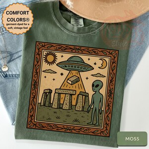 Op de afbeelding: Mosgroen T-shirt met een omlijnde afbeelding van een UFO, een alien en Stonehenge-achtige structuren. Het shirt heeft de tekst "COMFORT COLORS&reg; garment-dyed for a soft, vintage feel".