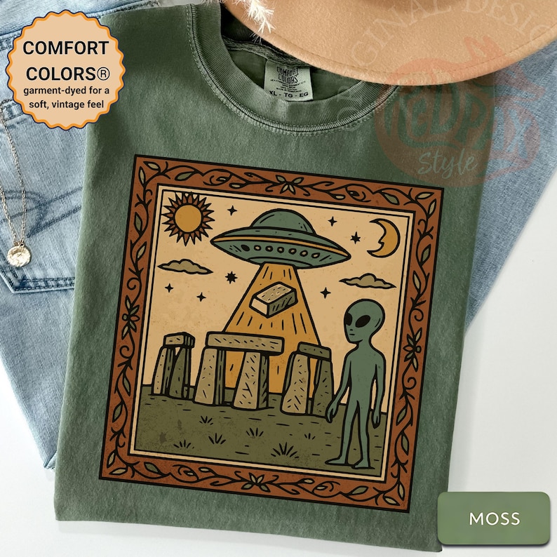 Op de afbeelding: Mosgroen T-shirt met een omlijnde afbeelding van een UFO, een alien en Stonehenge-achtige structuren. Het shirt heeft de tekst "COMFORT COLORS&reg; garment-dyed for a soft, vintage feel".