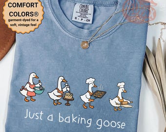 May include: Blue, garment-dyed t-shirt with a crew neck. The shirt features a whimsical illustration of geese baking, with the text "Just a baking goose." The shirt has a vintage feel and is labeled "Comfort Colors".