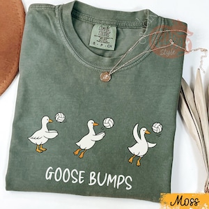以下が含まれることがあります： モスグリーンのTシャツ。3羽の白いガチョウがバレーボールをしているデザイン。白で「GOOSE BUMPS」の文字がプリントされています。金のネックレスがTシャツの上に置かれています。