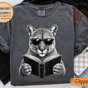 May include: A charcoal gray t-shirt featuring a graphic illustration of a mountain lion wearing black sunglasses and holding an open book. The shirt has a crew neckline and short sleeves. A circular badge on the top left reads COMFORT COLORS.