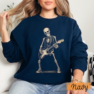 Peut inclure: Un sweat-shirt bleu marine &agrave; col rond avec une impression graphique repr&eacute;sentant un squelette portant un gilet et un n&oelig;ud papillon tout en jouant de la guitare &eacute;lectrique. Le squelette est beige clair, debout en position &eacute;cart&eacute;e avec des baskets.