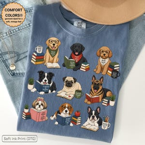 Puede incluir: Una camiseta azul con un estampado gráfico de nueve razas de perros diferentes, cada una representada con libros, gafas de lectura y pequeñas plantas en macetas. Los perros aparecen en varias posturas con pilas de libros coloridos y tazas. El texto incluye frases como Read More, Paws to Read, Book Lover, I'd Rather Be Reading, Good to Read, Cozy Books & Puppy Snugs, So Many Books So Little Time y Reading Fuels My Soul.