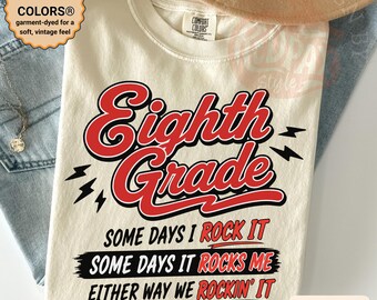Puede incluir: Camiseta de color marfil con la frase "Eighth Grade" en rojo y negro, junto con las frases "Some days I rock it" y "Some days it rocks me". La camiseta está teñida para una sensación suave y vintage.