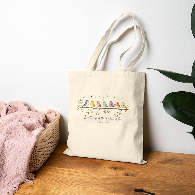 May include: A cream-colored canvas tote bag featuring a watercolor illustration of seven colorful birds perched on a leafy branch. Above the birds are musical notes, and below is the text: I will sing of the goodness of God, Psalm 89:1.