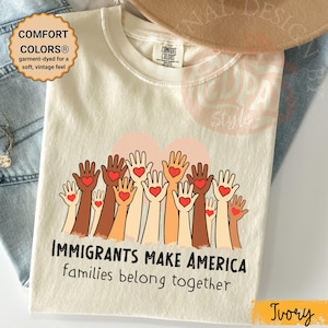 May include: Ivory-colored t-shirt with the text "IMMIGRANTS MAKE AMERICA families belong together." The shirt features a graphic of hands with hearts. The shirt is garment-dyed for a soft, vintage feel.