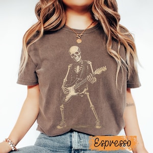 Peut inclure: Un t-shirt &agrave; manches courtes marron fonc&eacute; avec une impression graphique repr&eacute;sentant un squelette portant un gilet et jouant de la guitare &eacute;lectrique. Le squelette est repr&eacute;sent&eacute; debout, en couleur beige clair. Le mot Espresso est imprim&eacute; en dessous.