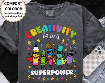 Könnte beinhalten: Dunkelgraues Sweatshirt mit dem Text "CREATIVITY is my SUPERPOWER" in bunten Farben. Das Design zeigt Bleistifte, einen Pinsel, ein Lineal und einen Marker als Superhelden. Das Sweatshirt hat einen Vintage-Look.