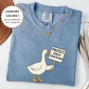 Puede incluir: Camiseta Comfort Colors azul claro con un pato de dibujos animados sosteniendo un cartel que dice "PROTECT PUBLIC EDUCATION". La camiseta tiene un tacto suave y vintage y una pequeña etiqueta Comfort Colors.