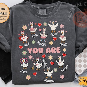 May include: Dark grey long-sleeved shirt featuring cartoon geese with hearts and flowers. The pink text reads "YOU ARE", with words like "brave", "kind", and "unique" around the geese. The shirt also has the text "COMFORT COLORS&reg;" on the upper left.