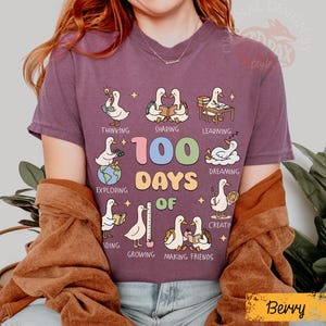 May include: Berry-colored t-shirt with a graphic featuring cartoon ducks engaged in various activities. The text "100 Days Of" is prominently displayed, with additional words like "Thinking," "Sharing," and "Learning." The shirt has a crew neck.