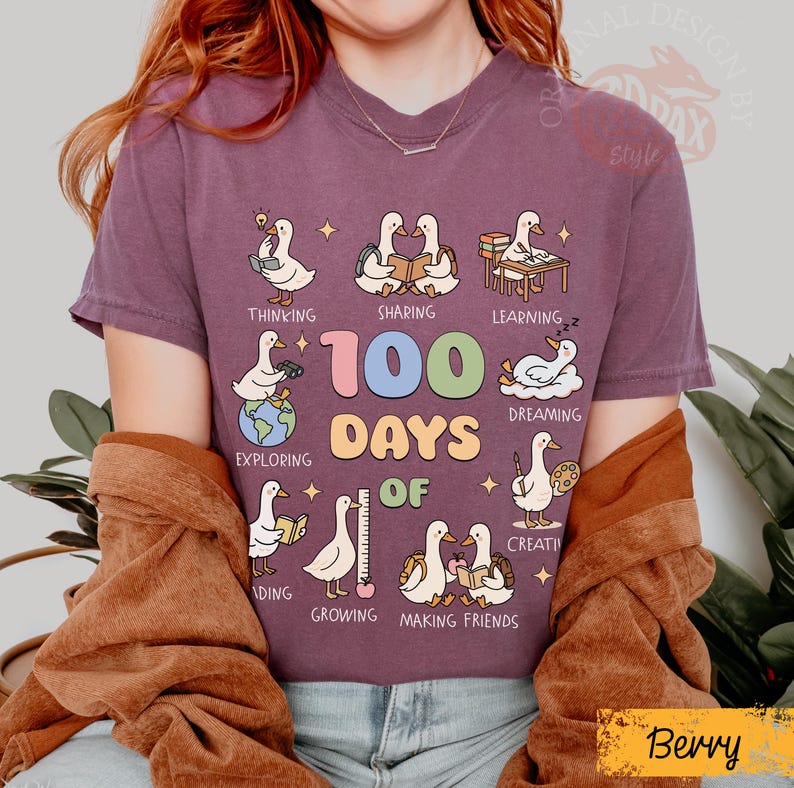 May include: Berry-colored t-shirt with a graphic featuring cartoon ducks engaged in various activities. The text "100 Days Of" is prominently displayed, with additional words like "Thinking," "Sharing," and "Learning." The shirt has a crew neck.
