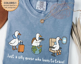 Könnte beinhalten: Hellblaues T-Shirt mit einer Grafik von drei Cartoon-Gänsen in Reisesituationen. Der Text "Just a silly goose who loves to travel" ist darunter gedruckt. Das T-Shirt hat einen weichen, Vintage-Look.