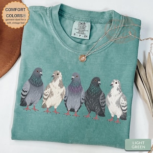 May include: A light green t-shirt featuring a graphic of five pigeons in various shades of gray and white. The shirt has a crew neckline and a Comfort Colors brand label on the inside collar. A thin silver chain necklace rests on the fabric.