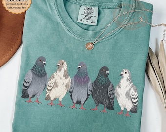 May include: A light green t-shirt featuring a graphic of five pigeons in various shades of gray and white. The shirt has a crew neckline and a Comfort Colors brand label on the inside collar. A thin silver chain necklace rests on the fabric.