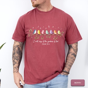May include: A berry-colored short-sleeve t-shirt featuring a graphic of seven colorful birds perched on a leafy branch with musical notes above them. Below the birds, the text reads, I will sing of the goodness of God, PSALM 89:1.