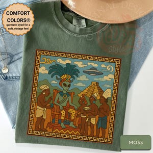 May include: Moss green t-shirt with a colorful graphic of an alien figure seated on a throne, surrounded by people, a pyramid, and a flying saucer. The shirt has the text "COMFORT COLORS® garment-dyed for a soft, vintage feel."