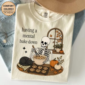 May include: A cream-colored t-shirt featuring a graphic of a skeleton whisking cookie dough in a bowl. The text reads having a mental bake down. A black cat sleeps nearby, with pumpkins, a cooling rack of cookies, and a window in the background.
