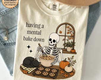Peut inclure: Un t-shirt crème avec une illustration de squelette fouettant de la pâte à biscuits dans un bol. Le texte indique having a mental bake down. Un chat noir dort à côté, avec des citrouilles, une grille de refroidissement et une fenêtre.