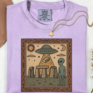 Op de afbeelding: Een lichtpaars Comfort Colors XL T-shirt met een gouden sterrenketting. Het shirt heeft een ingelijste afbeelding van een UFO, alien en Stonehenge-achtige structuren in vintage stijl.