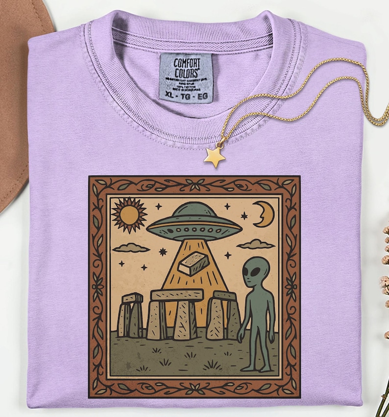 Op de afbeelding: Een lichtpaars Comfort Colors XL T-shirt met een gouden sterrenketting. Het shirt heeft een ingelijste afbeelding van een UFO, alien en Stonehenge-achtige structuren in vintage stijl.