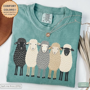 May include: A sage green t-shirt featuring a graphic print of five sheep in various colours including black, white, grey, and tan. The shirt has a crew neckline and a Comfort Colors brand label on the inside collar. The fabric has a vintage, garment-dyed appearance.