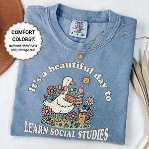 Puede incluir: Camiseta Comfort Colors azul claro con el texto "It's a beautiful day to learn social studies". El gráfico presenta un pato de dibujos animados sosteniendo libros y una bebida, rodeado de flores. Un collar dorado descansa sobre la camisa.