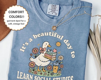 May include: A light blue Comfort Colors t-shirt with the text "It's a beautiful day to learn social studies." The graphic features a cartoon duck holding books and a drink, surrounded by flowers. A gold necklace rests on the shirt.