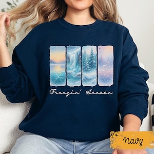 May include: Navy blue sweatshirt featuring a winter landscape design. The design includes four rectangular panels depicting snowy scenes with a waterfall, trees, and frozen lake. The words "Freezin' Season" are written in cursive below the panels.