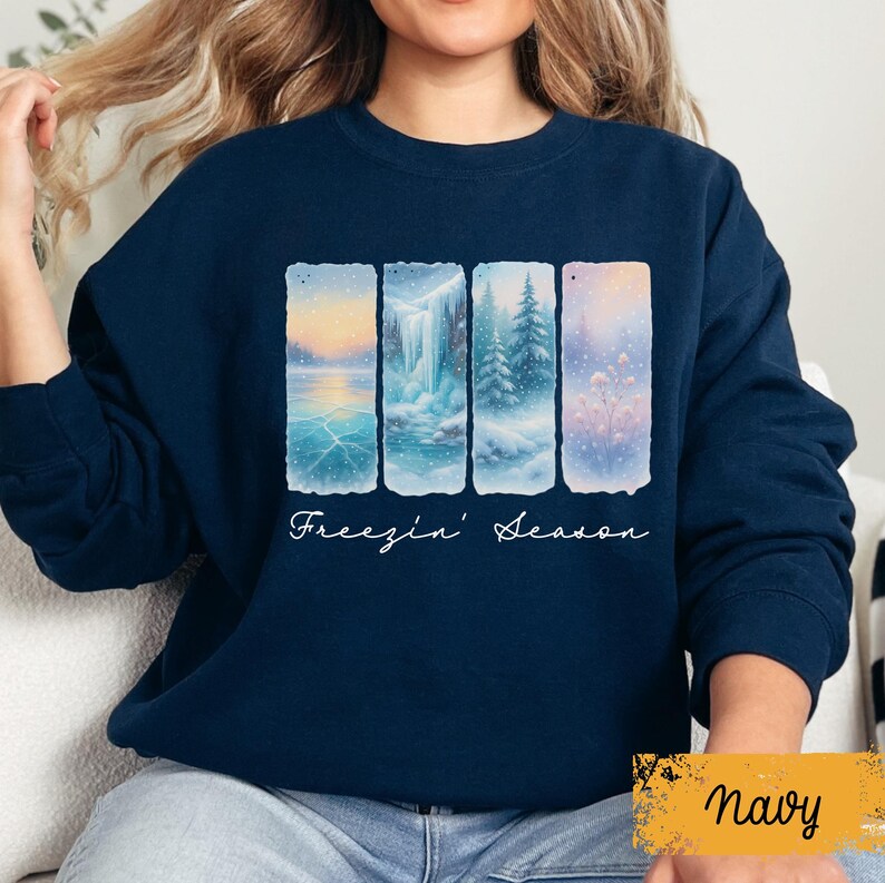 May include: Navy blue sweatshirt featuring a winter landscape design. The design includes four rectangular panels depicting snowy scenes with a waterfall, trees, and frozen lake. The words "Freezin' Season" are written in cursive below the panels.