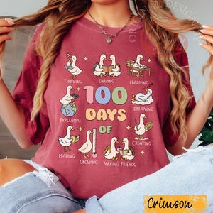 May include: Crimson-colored t-shirt with a colorful graphic celebrating "100 Days Of" learning. The design features cartoon geese engaged in various activities like reading, sharing, and exploring. The text "Crimson" is at the bottom.