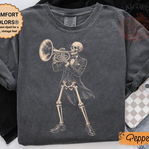 May include: A charcoal grey short-sleeve t-shirt featuring a graphic of a skeleton wearing a tuxedo and playing a trumpet. The shirt has a crew neckline and a vintage-washed appearance. The text COMFORT COLORS is visible on the inner neck label.