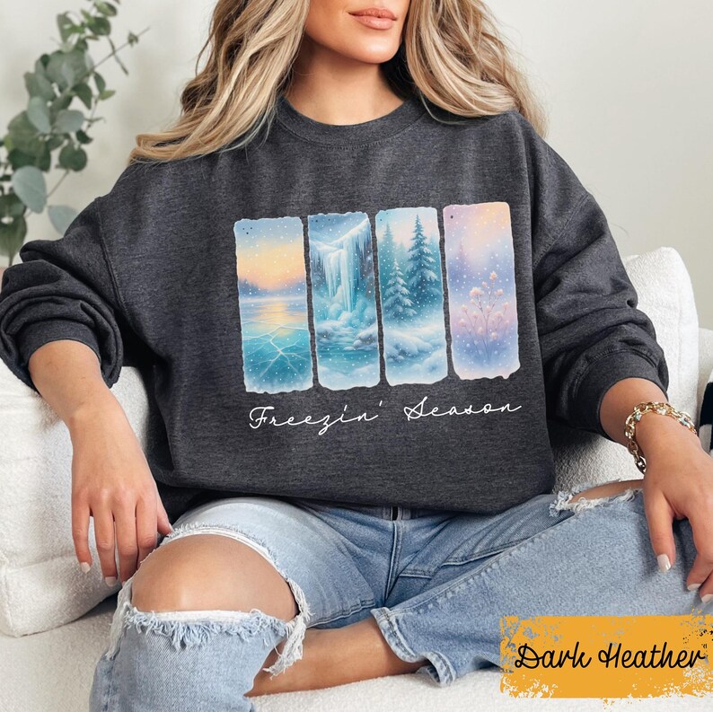 May include: Dark heather gray sweatshirt featuring a winter landscape design with the text "Freezin' Season". The graphic shows four rectangular panels depicting a frozen lake, waterfall, snowy trees, and a field of snow. The sweatshirt is a comfortable, casual style.