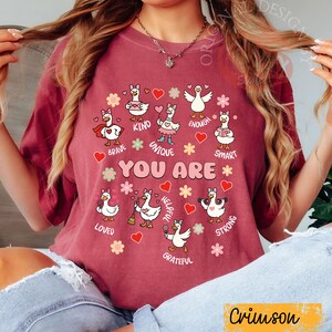 May include: Crimson t-shirt with a design of cartoon geese, each representing a positive trait such as "Brave," "Kind," and "Unique." The phrase "YOU ARE" is central, with hearts and flowers. The shirt's colour is described as "Crimson."