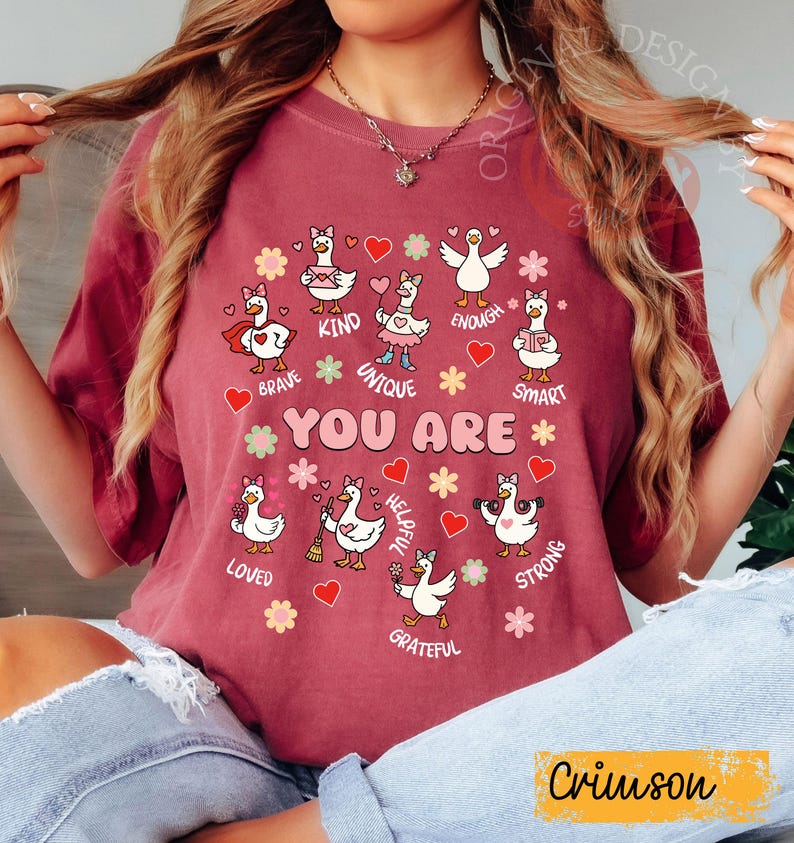 May include: Crimson t-shirt with a design of cartoon geese, each representing a positive trait such as "Brave," "Kind," and "Unique." The phrase "YOU ARE" is central, with hearts and flowers. The shirt's colour is described as "Crimson."