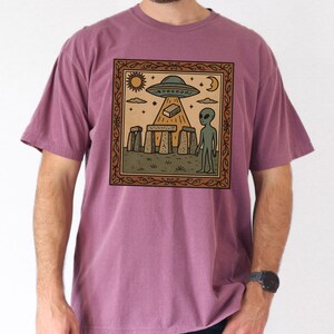 Op de afbeelding: Een mauvekleurig T-shirt met een ingelijste afbeelding van een buitenaardse sc&egrave;ne. De afbeelding toont een UFO, een alien en Stonehenge. Het ontwerp bevat een zon, maan en sterren. Het shirt is gemaakt van een zacht materiaal.
