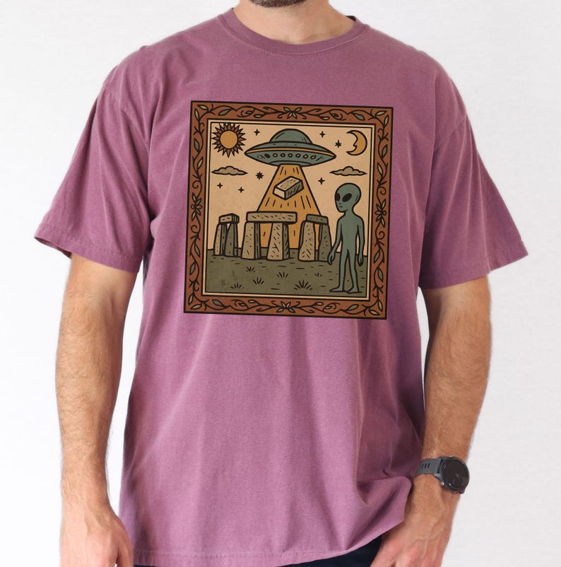 Op de afbeelding: Een mauvekleurig T-shirt met een ingelijste afbeelding van een buitenaardse sc&egrave;ne. De afbeelding toont een UFO, een alien en Stonehenge. Het ontwerp bevat een zon, maan en sterren. Het shirt is gemaakt van een zacht materiaal.
