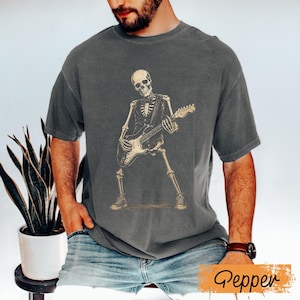 Peut inclure: Un t-shirt gris anthracite &agrave; manches courtes avec une impression graphique repr&eacute;sentant un squelette portant un gilet et jouant de la guitare &eacute;lectrique. Le squelette est repr&eacute;sent&eacute; debout, portant des baskets, dans des tons beige clair.