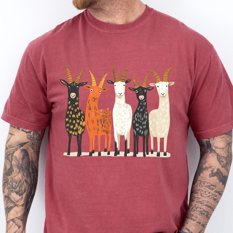 May include: A maroon t-shirt featuring a graphic of five stylized goats in various colors: black, orange, white, and cream. The goats stand side-by-side, each with detailed horns and a unique pattern. The shirt is made of a soft material.