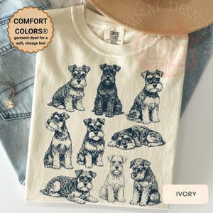 Peut inclure: T-shirt ivoire avec plusieurs illustrations de chiens Schnauzer dans diverses poses. Le t-shirt porte le texte "COMFORT COLORS® garment-dyed for a soft, vintage feel". Le t-shirt est fait d'une matière douce.