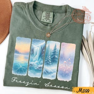 Puede incluir: Camiseta verde musgo con un dise&ntilde;o de paisaje invernal. El dise&ntilde;o presenta cuatro paneles rectangulares que representan un lago helado, una cascada, &aacute;rboles nevados y flores. El texto "Freezin' Season" est&aacute; impreso debajo.