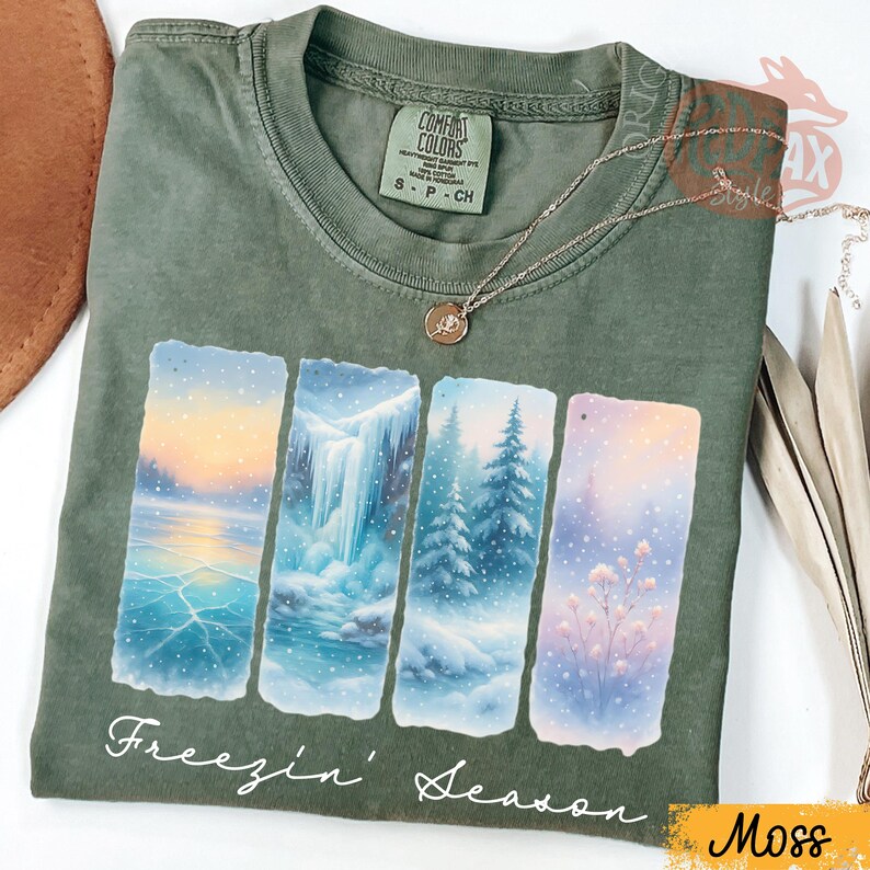 Puede incluir: Camiseta verde musgo con un dise&ntilde;o de paisaje invernal. El dise&ntilde;o presenta cuatro paneles rectangulares que representan un lago helado, una cascada, &aacute;rboles nevados y flores. El texto "Freezin' Season" est&aacute; impreso debajo.