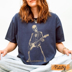 Peut inclure: Un t-shirt bleu marine &agrave; manches courtes avec une impression graphique repr&eacute;sentant un squelette portant un gilet et un n&oelig;ud papillon tout en jouant de la guitare &eacute;lectrique. Le squelette est en couleur beige clair, dans une pose dynamique.