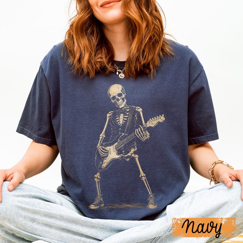 Peut inclure: Un t-shirt bleu marine &agrave; manches courtes avec une impression graphique repr&eacute;sentant un squelette portant un gilet et un n&oelig;ud papillon tout en jouant de la guitare &eacute;lectrique. Le squelette est en couleur beige clair, dans une pose dynamique.
