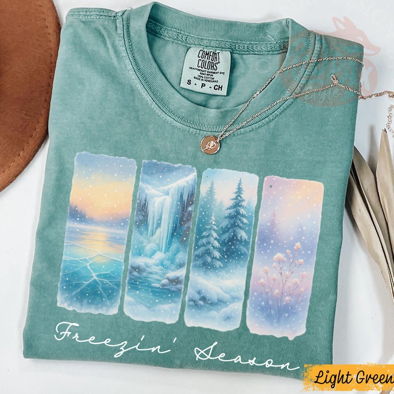 Puede incluir: Camiseta verde claro Comfort Colors con un gr&aacute;fico de tem&aacute;tica invernal. El gr&aacute;fico presenta cuatro paneles rectangulares que representan escenas invernales, incluyendo un lago helado, una cascada y &aacute;rboles nevados. El texto "Freezin' Season" est&aacute; impreso en la parte inferior.