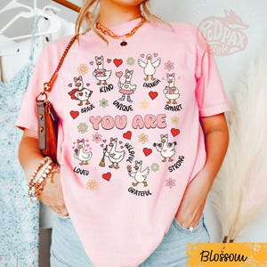 May include: A pink t-shirt with the words "YOU ARE" and illustrations of geese with positive affirmations. The shirt features the word "Blossom" in a yellow rectangle. The geese are surrounded by hearts and flowers.